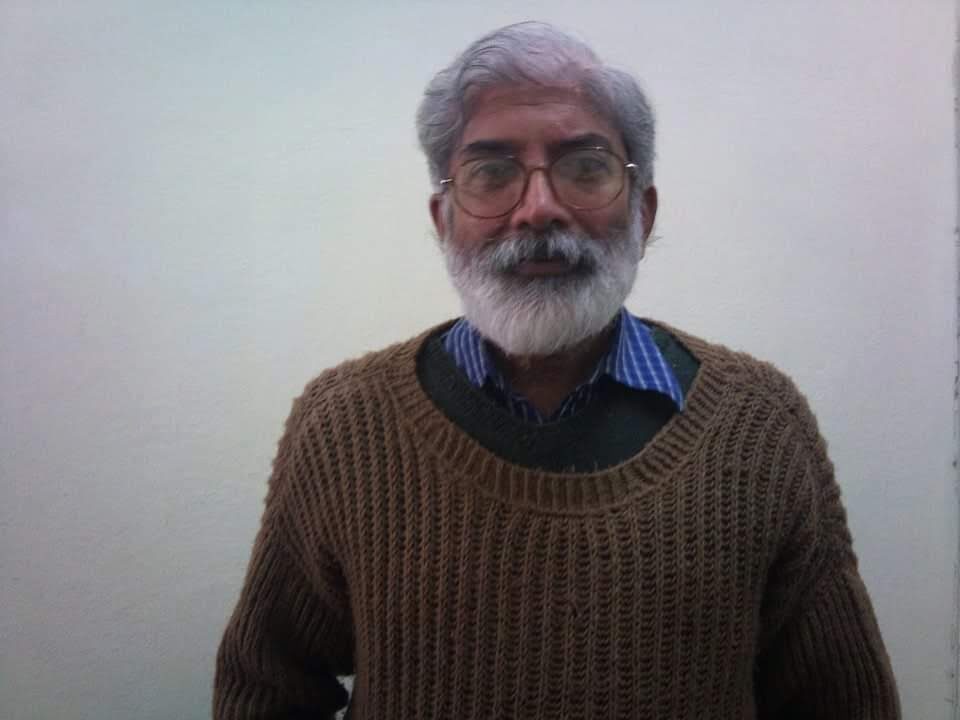 An older man with a grey beard and rimmed spectacles in a brown knit sweater, looking straight at the camera against a plain wall