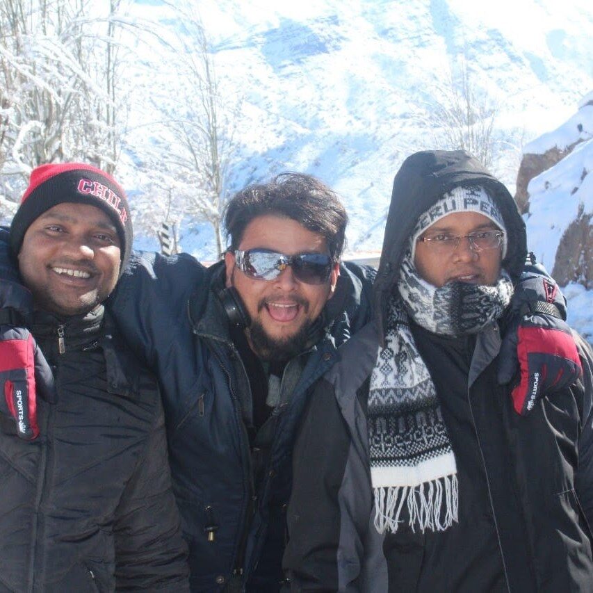 Three friends in winter jackets and beanies on a snowy mountainside, the one in the middle wearing sunglasses and sticking his tongue out