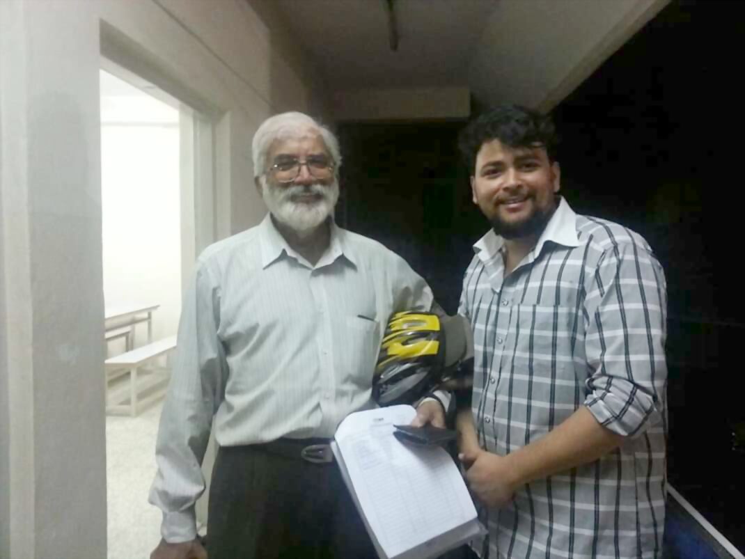An older man with a white beard and glasses holding a black and yellow helmet and a printed sheet, standing beside a younger man in a plaid shirt in a corridor