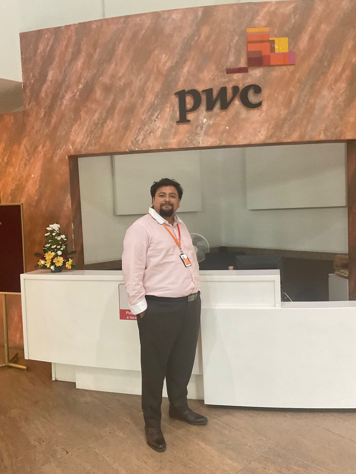 A man in a light pink shirt with rolled cuffs and an ID badge on a lanyard, standing in front of a reception desk with a company logo on the wall